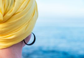 Fotografia colorida. Na imagem, uma mulher com lenço amarelo na cabeça aparece olhando para o mar. Ela usa um brinco preto de argola.