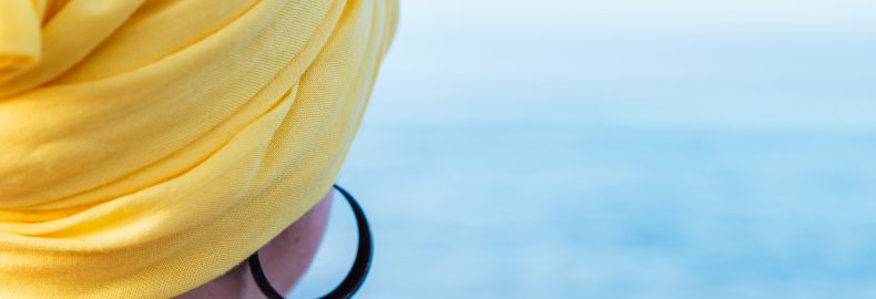 Fotografia colorida. Na imagem, uma mulher com lenço amarelo na cabeça aparece olhando para o mar. Ela usa um brinco preto de argola.