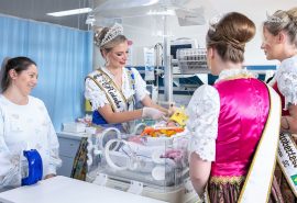 Fotografia colorida. Na imagem, a mãe da paciente Eloá, as princesas e a rainha da 37ª Oktoberfest aparecem olhando para paciente, que aparece deitada em uma incubadora com a faixa de rainha.