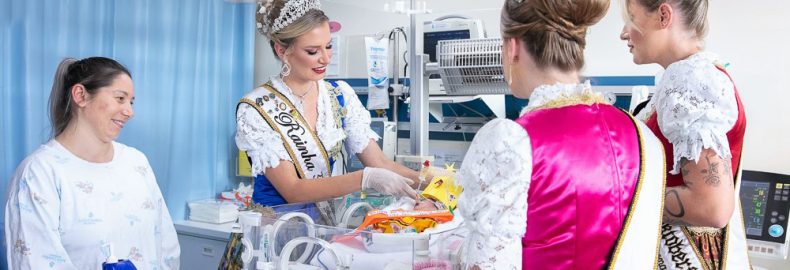 Fotografia colorida. Na imagem, a mãe da paciente Eloá, as princesas e a rainha da 37ª Oktoberfest aparecem olhando para paciente, que aparece deitada em uma incubadora com a faixa de rainha.