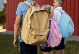 Fotografia. Três crianças voltando da escola com suas respectivas mochilas nas cores amarelo, rosa e azul.