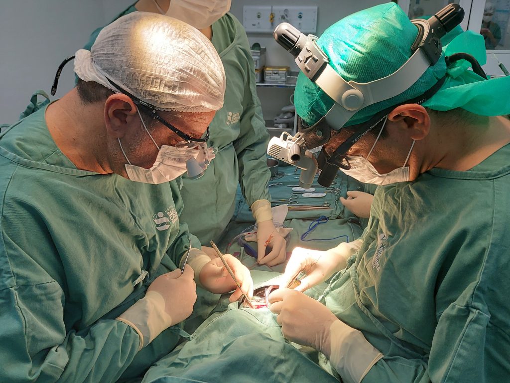 Na imagem aparecem médicos paramentados realizando a cirurgia de cardiopatia congênita em um  bebê de quatro meses.
